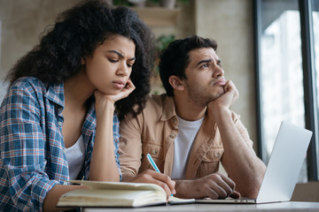 Pensive students studying together, learning language, exam preparation, taking notes in library. Tired mixed race colleagues freelancers working project, using laptop, brainstorming at workplace