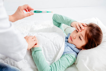 Sick child with flu fever laying in bed and mother holding thermometer