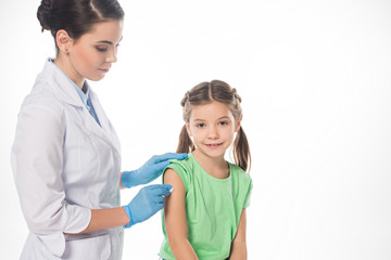 Female pediatrician with cotton wool rubbing shoulder of smiling kid isolated on white