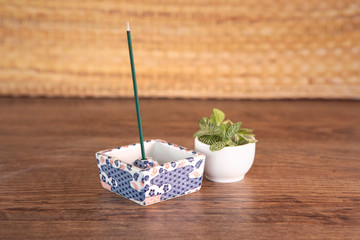 Composition with japanese style incense stick burning in beautiful ceramic incense stand, wooden table background, some fresh green plant.