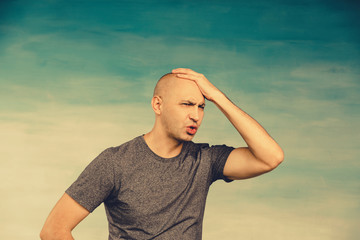 A bald man in a gray t-shirt holds his hand with head as in a severe headache in frustration on a blue background.