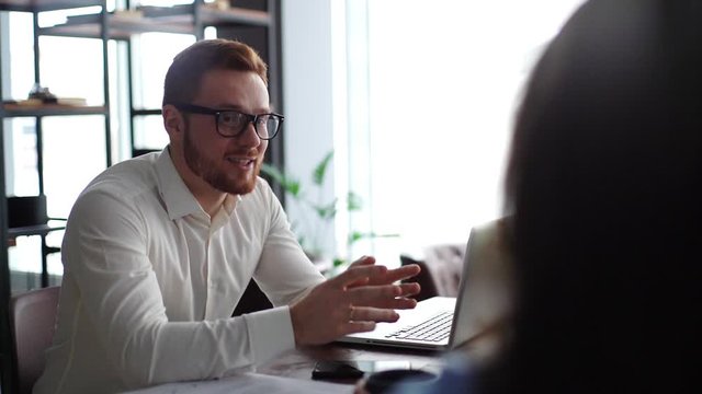 Handsome male manager with glasses and beard is consulting client, explaining deal details convincing buy services in modern office meeting against large window. Shooting in slow motion.