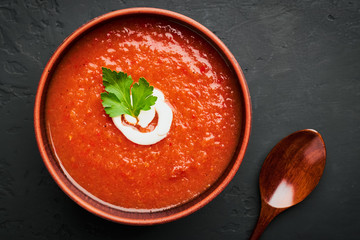 Top view close-up of a brown bowl of tomato soup with parsley and milk cream and a wooden spoon on a dark background