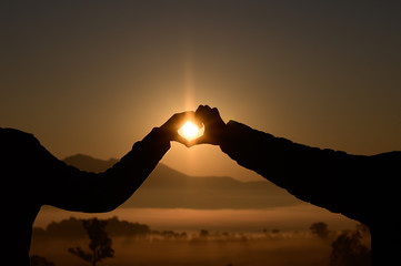 Silhouette hand in heart shape with sunrise in the middle and mountain background.