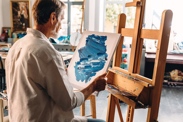 Back view of male painter holding a canvas with abstract picture sitting at easel