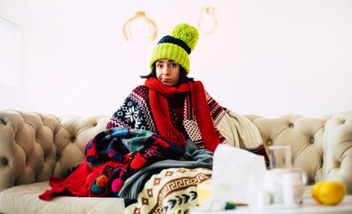 Caught cold. Cute young ill woman is feeling cold and sick sitting on a couch and being warmly dressed in a sweater, scarf and green knitted hat.
