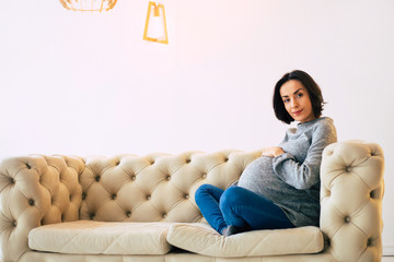 Obraz premium Future mom. Young pregnant mom in casual clothes is sitting on a couch at home in a lotus position and looking in the camera.