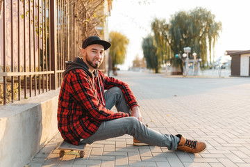 Skateboarding. A handsome guy with a beard, in a cap and fashionable clothes poses with a skate on...