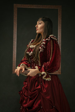 Proud. Portrait of medieval young woman in red vintage clothing standing on dark background. Female model as a duchess, royal person. Concept of comparison of eras, modern, fashion, beauty.