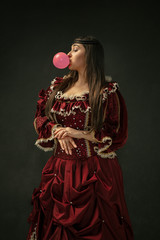 Pink bubble gum. Portrait of medieval young woman in red vintage clothing standing on dark...