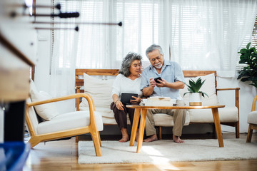 Happy Asian senior couple using smartphone together on couch in living room, older man texting message or chatting with family, elderly woman watching interested on gadget , Aging society concept