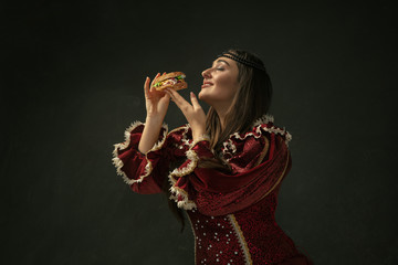 Passioned. Portrait of medieval young woman in red vintage clothing eating burger on dark background. Female model as a duchess, royal person. Concept of comparison of eras, modern, fashion, beauty.