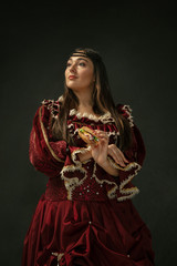 Fototapeta premium First love. Portrait of medieval young woman in red vintage clothing holding burger on dark background. Female model as a duchess, royal person. Concept of comparison of eras, modern, fashion, beauty.