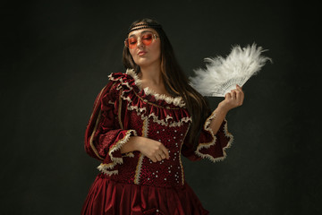 Fototapeta premium Red eyewear and fluffy fan. Portrait of medieval young woman in red vintage clothing on dark background. Female model as a duchess, royal person. Concept of comparison of eras, modern, fashion, beauty