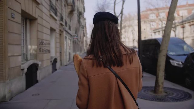 Camera follows beautiful elegant happy woman walking along Paris street with paper shoppping food bag slow motion.