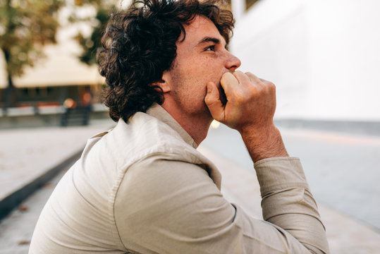 Side view of young man with curly hair looking at one side, sitting outside. Portrait of handsome male with curly hair has pensive expression, take a rest in the city street.