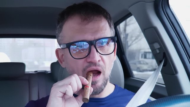 Caucasian man behind steering wheel pulls out cigar and sticks it in his mouth