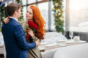 elegant man and attractive redhaired lady in dress on a date in restaurant, gallant good-looking male in tux brought red roses for lady in a sign of attention. love, dating, couple, romantic concept