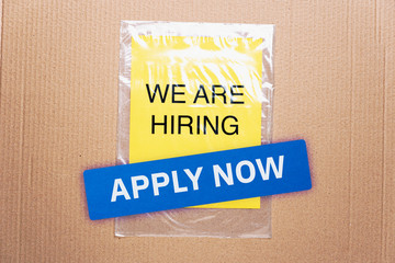 Apply now job button here hiring resources register online concept. We are hiring printed on yellow paper on transparent plastic bag with cardboard background.
