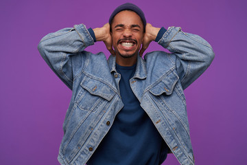 Studio shot of young stressed dark skinned bearded guy frowning his face with closed eyes while covering ears with raised palms, avoiding loud sounds, isolated over purple background