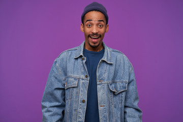Open-eyed young beautiful dark haired bearded male in blue cap, pullover and jeans coat raising amazedly eyebrows while looking joyfully at camera, standing over purple background