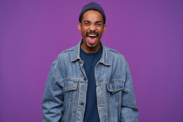 Indoor photo of young angry dark skinned brunette male with beard frowning his face while screaming fiercely with wide mouth opened, isolated over purple background