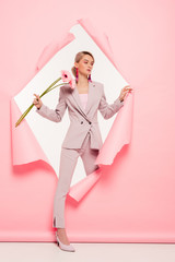 attractive girl in trendy suit holding flowers and posing in torn paper, on white