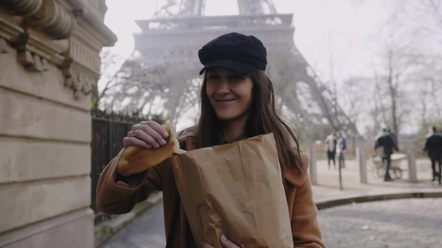 Beautiful happy European woman walking near Eiffel Tower, giving a piece of French baguette to camera man slow motion.