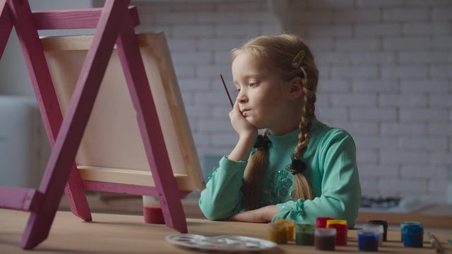 Dreamy cute preadolescent girl with paintbrush pondering over future painting, looking for inspiration and fresh ideas, fully engrossed in creativity while sitting near easel in domestic kitchen.