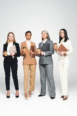 Fototapeta na wymiar four multicultural businesswomen holding folders and paper cups while standing on white