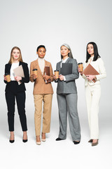 Obraz premium four multicultural businesswomen holding folders and paper cups while standing on white
