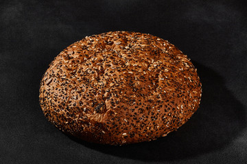 Fresh, tasty baked round bread sprinkled with black sesame and sunflower seeds against black background with copy space. Close-up