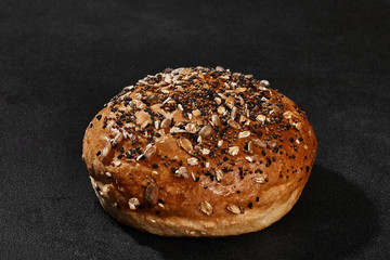 Fresh, delicious baked bun sprinkled with sesame and sunflower seeds against black background with copy space. Rural cuisine or bakery. Close-up