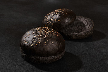 Fresh, baked, whole and cut in half black or chocolate bun sprinkled with sesame seeds against black background with copy space. Close-up