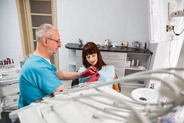 Obraz premium Senior man doctor dentist consulting young pretty woman patient holding artificial jaw at the modern dental office