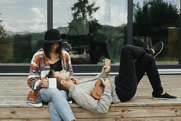 Hipster man playing on ukulele for his beautiful stylish woman, relaxing on wooden porch on...