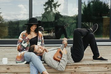 Stylish hipster couple sitting on wooden porch, relaxing with ukulele  on background of modern...