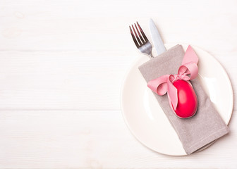 Easter composition. Creative table setting for the holiday of Easter. Red easter egg on the gray napkin and white plate, fork and knife on the white wooden background. Copy space, top view