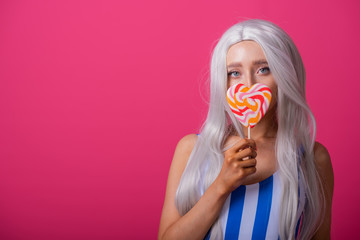 Portrait of a slender woman in a blonde wig posing with a lollipop on a pink background. Cute girl...