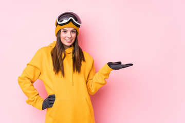 Young caucasian skier woman isolated showing a copy space on a palm and holding another hand on waist.
