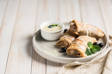 A few pancakes with filling on a gray plate with blueberries and mint, with condensed sweet milk in a gravy boat on a white wooden background.