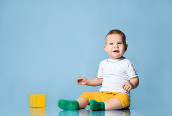 Smiling infant baby boy toddler in yellow pants is sitting on the floor near yellow cube, looking up