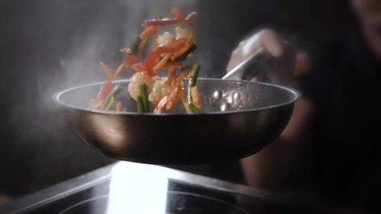 Cook in transparent sterile gloves in the kitchen throws up a hot frying wok with chicken, vegetables and noodles in a black pan