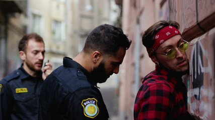Police arrest. View of young man drug dealer near the wall being handcuffed. Policeman seacrhes an...