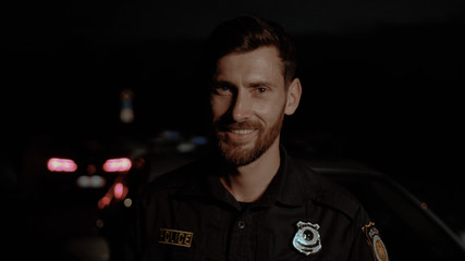 Portrait of cheerful handsome policeman smiling broadly against his patrol car at night. Police officers on durty in the city. © Fractal Pictures