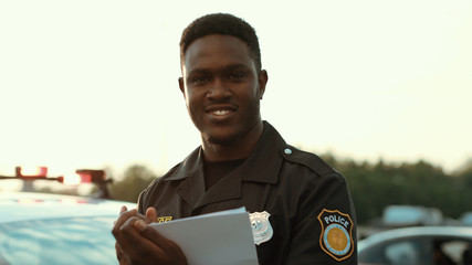 Portrait of cheerful smiling policeman holding papers leaning on the police car posing at sunlight....