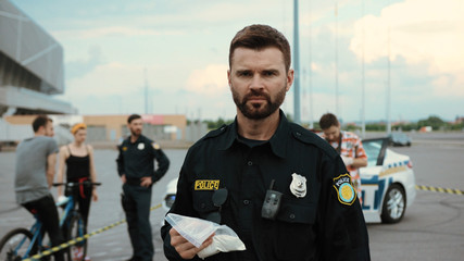 Portrait of concentrated police officer examining illegal drugs package looking straight performing investigation in the street. Police patrol in action.