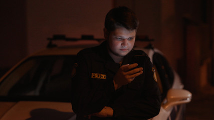 Portrait handsome young police officer chatting in social network on mobile phone leaning on the police car hood in the street at night.