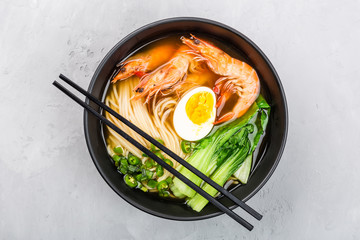 Asian noodle soup, ramen with shrimp, vegetables and egg in black bowl on gray concrete background. Flat lay, Top view, mockup, overhead. Healthy food concept