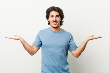 Young handsome man against a white background makes scale with arms, feels happy and confident.